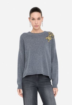 Maglione grigio lavorato a maglia con una vestibilità ampia, scollatura rotonda e decorazioni in giallo e oro sulla spalla. Abbinato a jeans scuri.