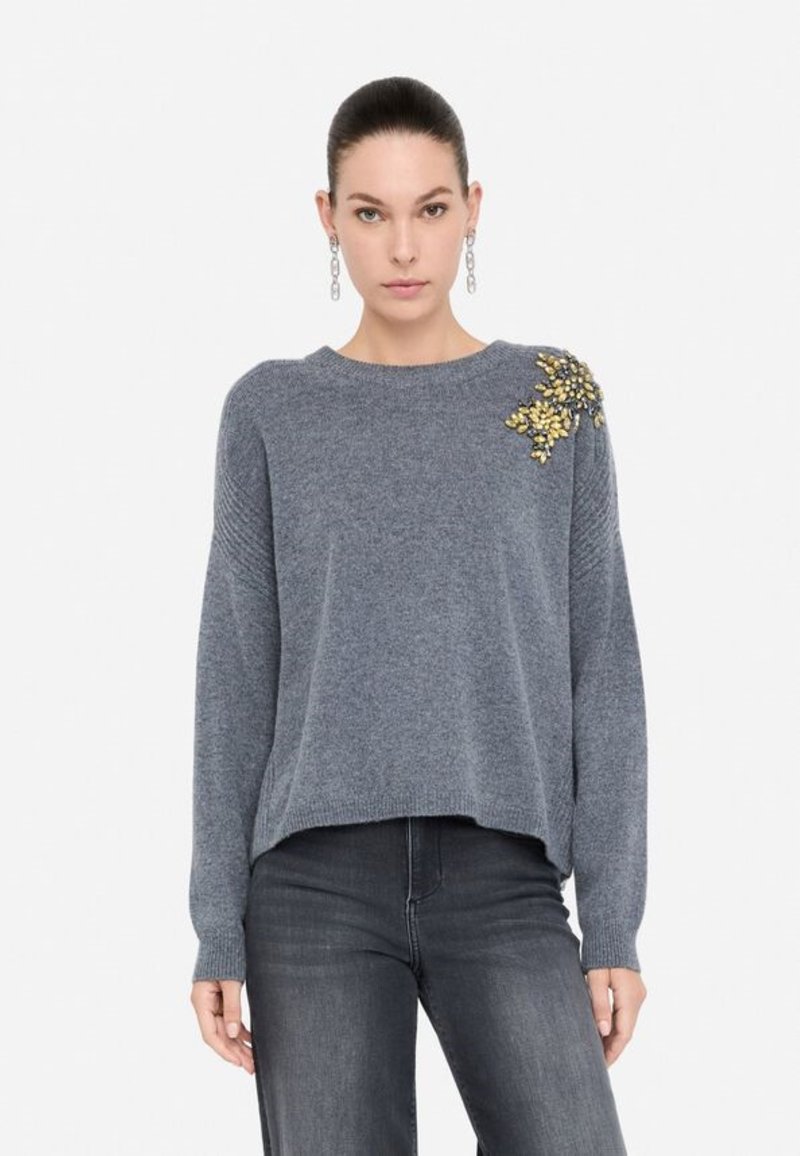 Maglione grigio lavorato a maglia con una vestibilità ampia, scollatura rotonda e decorazioni in giallo e oro sulla spalla. Abbinato a jeans scuri.