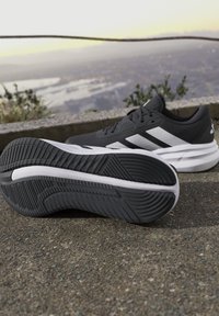 Chaussures de sport noir et blanc sur le trottoir montrant le motif de la semelle, avec un paysage extérieur flou et une rambarde en arrière-plan.
