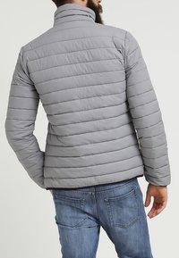 Uomo che indossa un giubbotto imbottito grigio chiaro e jeans blue, visto da dietro contro uno sfondo chiaro e semplice.