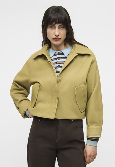 Γυναίκα με σκούρα μαλλιά που φοράει ένα κοντό μουσταρδί μπουφάν πάνω από ένα ριγέ μπλε πουκάμισο με γιακά και σκούρο παντελόνι, όρθια μπροστά σε απλό φόντο.