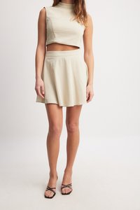 Costum în două piese de culoare bej deschis; top scurt fără mâneci cu guler înalt și fustă scurtă evazată. Material texturat, design minimalistic.