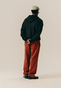 Άνδρας φορώντας μαύρο πουλόβερ, κόκκινα παν trousers, μπεζ καπέλο και μαύρα παπούτσια στέκεται με τα χέρια δεμένα πίσω από την πλάτη, γυρισμένος προς τα πίσω σε απλό φόντο.