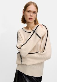 Suéter de canalé crema con costuras en negro contrastantes, escote barco y mangas oversize. El material parece suave con un acabado texturizado.
