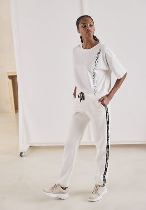 Weißes Baumwoll-T-Shirt mit vertikal in Schwarz bedrucktem "FRENCH CONNECTION", kombiniert mit cremefarbenen Jogginghosen mit schwarzen Seitenstreifen und Kordelzug.