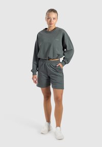 Felpa verde scuro con vestibilità ampia e orlo elastico, abbinata a pantaloni corti coordinati con vita elastica e tasche laterali. Sneakers bianche.