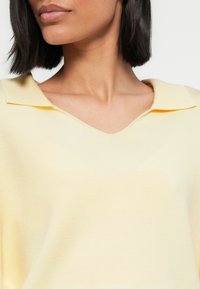 Ανοιχτό κίτρινο πουλόβερ με λαιμόκοψη σε σχήμα V και επίπεδη, διπλωμένη γιακά. Η υφή του πλεκτού είναι λεία με διακριτική γυαλάδα.