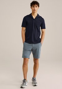 Mornarska polo majica s kratkimi rokavi in žepom na prsih, v kombinaciji s svetlo modrimi kratkimi hlačami. Model nosi sive superge. Preprost, sproščen dizajn.