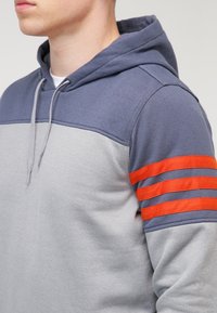 Mikina s kapucí šedého těla, tmavě modré horní části a oranžovým pruhovaným akcentem na rukávu. Měkká textura, design na přehození.