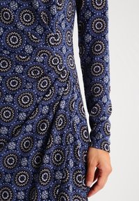 Long-sleeve dress with a deep blue pattern featuring circular motifs in black and white, displaying a smooth texture and draped fabric.