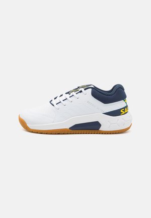 Scarpa atletica bianca con accenti blu navy, tomaia in rete testurizzata e suola in gomma. Presenta lacci e un'etichetta sul tallone di colore giallo.