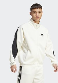 Veste de sport blanche avec des accents noirs sur les manches, col haut et fermeture éclair à l'avant. Présente des poignets et un ourlet côtelés, ainsi qu'un petit logo sur la poitrine.