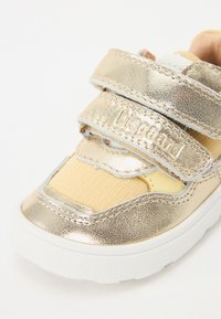Baskets métalliques dorées avec des panneaux en tissu beige clair, dotées de deux bandes Velcro réglables et d'une semelle en caoutchouc blanche.