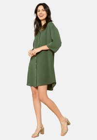 Green button-up dress with three-quarter sleeves, a curved hem, and a soft texture. Worn with gold heeled sandals.
