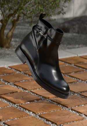 Black leather ankle boot with side strap and silver logo detail, floating above wooden tile floor with gray pebbles and blurred plant background.