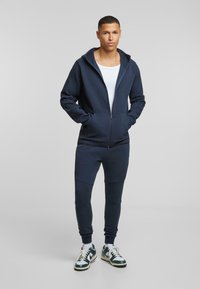 Námornícka mikina na zips s vyhovujúcimi teplákmi, bavlnený materiál, žebrované manžety a pás, obsahuje predné vrecká, skombinované so zelenými a bielymi teniskami.