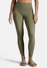 Olivgrüne, hochtaillierte Leggings mit einer glatten, dehnbaren Textur und flachen Nähten. Kombiniert mit hellbeigen Rutschesandalen.