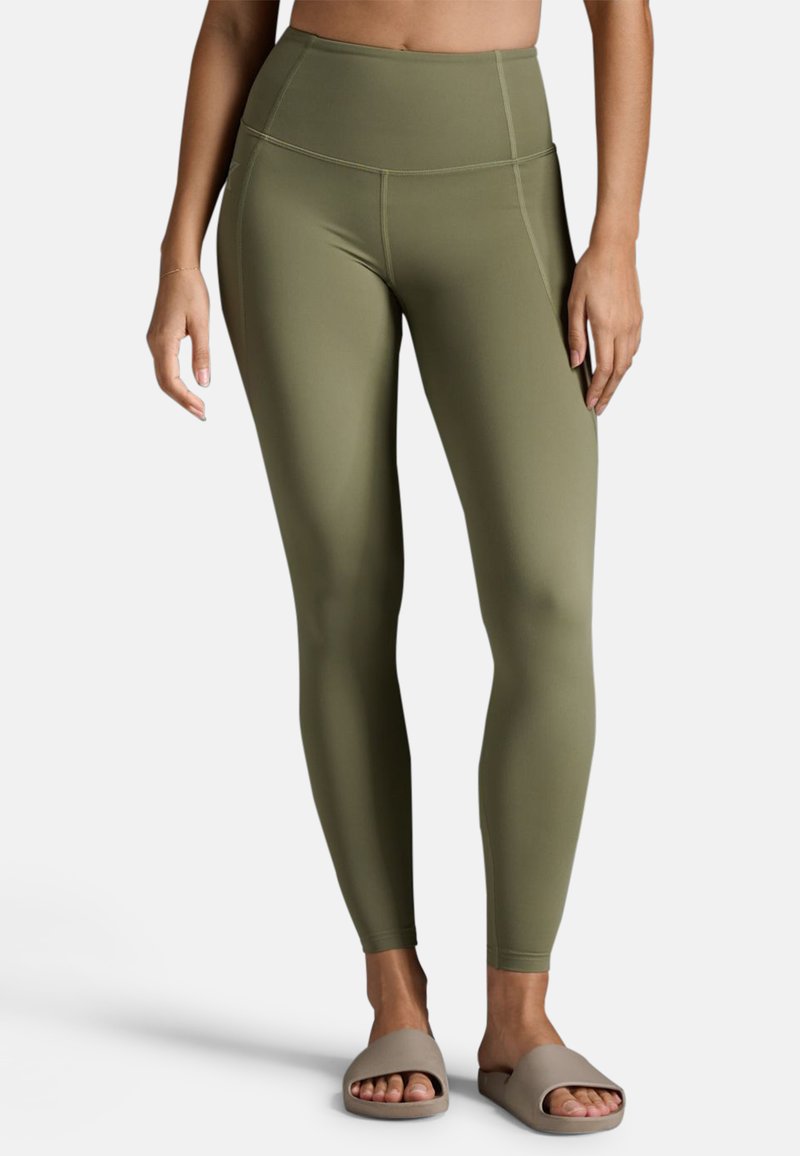 Olivgrüne, hochtaillierte Leggings mit einer glatten, dehnbaren Textur und flachen Nähten. Kombiniert mit hellbeigen Rutschesandalen.