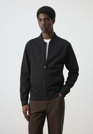 Joven con chaqueta negra con cremallera y pantalones marrones, de pie con una mano en el bolsillo frente a un fondo claro y liso.
