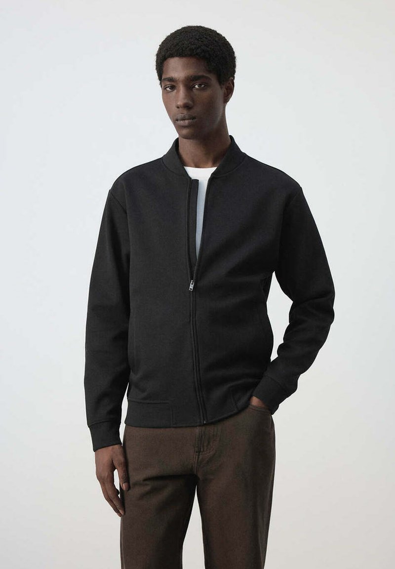 Jeune homme portant une veste noire zippée et un pantalon marron, debout avec une main dans la poche devant un fond clair uni.