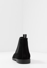 Botín de ante negro con paneles laterales elásticos, suela plana negra y una lengüeta trasera con rayas negras y grises.