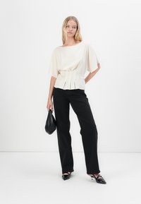 Top plissettato color crema con maniche ampie, abbinato a jeans neri a vita alta. Scarpe nere a punta e una piccola borsa nera completano il look.