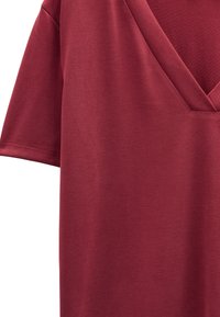 Burgundowa koszulka z dekoltem w serek i krótkim rękawem, wykonana z gładkiego, lekkiego materiału. Charakteryzuje się prostym, czystym wzornictwem bez widocznych wzorów lub elementów metalowych.