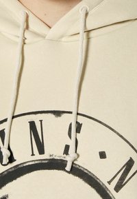 Sudadera crema con un diseño de logo redondo que presenta texto negro. Tiene una textura suave y cordones blancos con extremos anudados.
