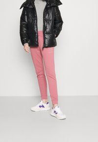 Černá lesklá péřová bunda s kapucí, šedý svetr, růžové tepláky s logem a bílé tenisky s fialovými akcenty. Neformální outfit.