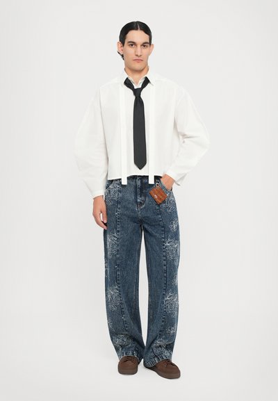 Camicia bianca a maniche lunghe con cravatta nera, abbinata a jeans larghi in denim blu con ricami floreali. Scarpe marroni completano l'outfit.