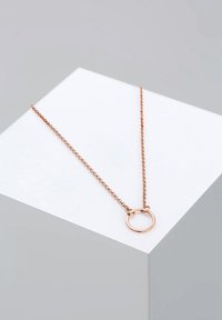 Rose gold halskæde med et minimalistisk design, der har en cirkulær vedhæng hængende fra en fin kæde, præsenteret på en geometrisk overflade.