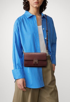 Burgundy leather crossbody bag with gold hardware, featuring a logo on the front. Worn with a blue striped shirt and beige trousers.