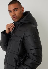 Sort puffjakke med hætte, der har en blank finish, knaplukning foran og en brystlomme. Tekstureret stof med synlige sømme.
