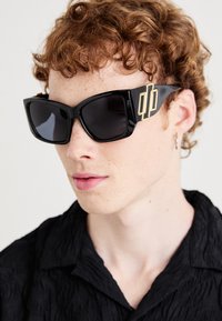 Occhiali da sole neri oversize con una finitura lucida, caratterizzati da un accento geometrico dorato sulla stanghetta. Indossati da una persona con capelli ricci.
