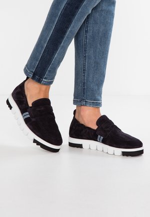 Mocassins en daim bleu marine avec une semelle blanche flexible et des accents noirs ; dotés de deux rayures bleues sur le devant. Portés avec un jean slim.