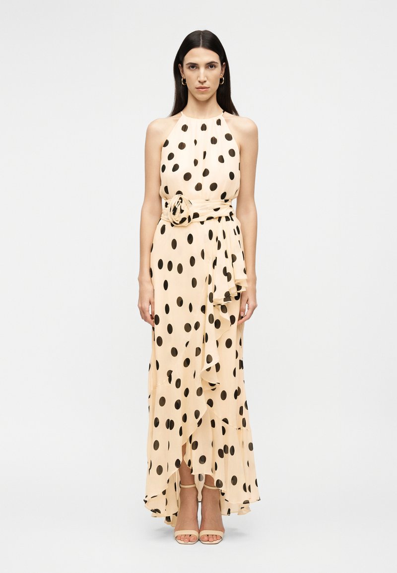 Γυναίκα με μπεζ αμάνικο φόρεμα με μαύρες πουά βούλες, ζώνη που ταιριάζει και ανοιχτά τακούνια, στέκεται μπροστά σε απλό φόντο.