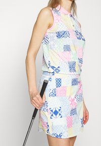 Färgglad golfoutfit med en ärmlös skjorta i lapptäcksmönster i pastellfärger och en matchande kjol, hållandes en golfklubba.