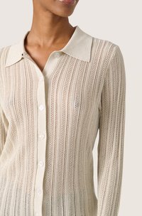 Cardigan a bottone color crema lavorato a maglia con colletto. Presenta strisce verticali e un motivo di punti aperti, evidenziando un design testurizzato.