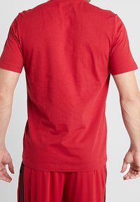 Muž viděný zezadu, oblečený v obyčejném červeném tričku a červených sportovních šortkách s černými pruhy, stojící na neutrálním pozadí.