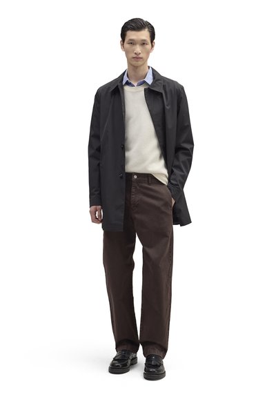 Manteau imperméable noir porté sur un pull crème et une chemise bleue à col, associé à un pantalon marron et des chaussures noires. Design simple et minimaliste.