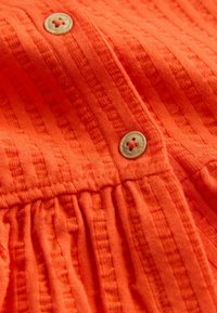 Oranssi teksturoidulla kankaalla, jossa pystysuorat uurteet ja vaaleanruskeat napit, jotka on kiinnitetty oranssilla langalla.