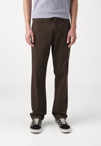 Braune Baumwollhosen mit geradem Schnitt, ausgestattet mit einem Frontknopfverschluss und Taschen. Kombiniert mit gemusterten schwarz-weißen Sneakers.