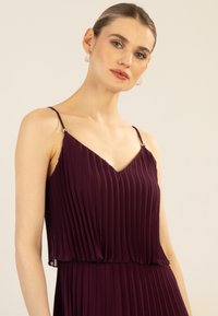 Lila plissiertes Kleid mit dünnen Spaghettiträgern und V-Ausschnitt, das metallische Akzente an den Trägern aufweist. Weiche, fließende Stoffstruktur.