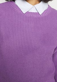 Fioletowy sweter dziany o teksturowanej tkaninie, z okrągłym dekoltem i widocznymi prążkowanymi detalami na mankietach oraz na dole.