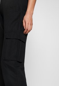 Svarta cargobyxor med stora sidofickor. Tillverkade av slitstarkt tyg, med en lös passform och synliga sömnadsdetaljer längs sömmarna.