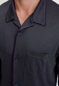 Donkergrijze shirt met een knoopsluiting, contrasterende bordeauxrode piping op de kraag en een borstzak. Zachte textuur en stof.