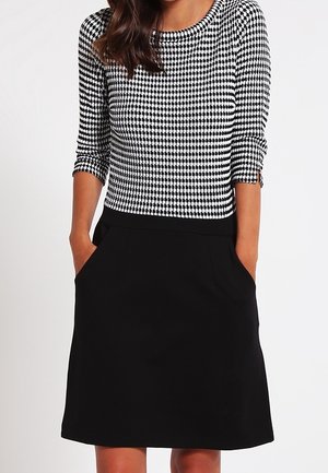 Frau trägt ein schwarz-weiß gemustertes Oberteil mit Hahnentritt und Dreiviertelärmeln sowie einen einfarbigen schwarzen knielangen Rock mit Taschen.