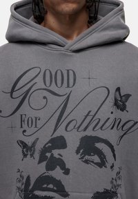 Felpa grigia con cappuccio caratterizzata da una grande grafica con il testo "GOOD For Nothing" e dettagli a forma di farfalla, che presenta una vestibilità rilassata e una texture morbida.