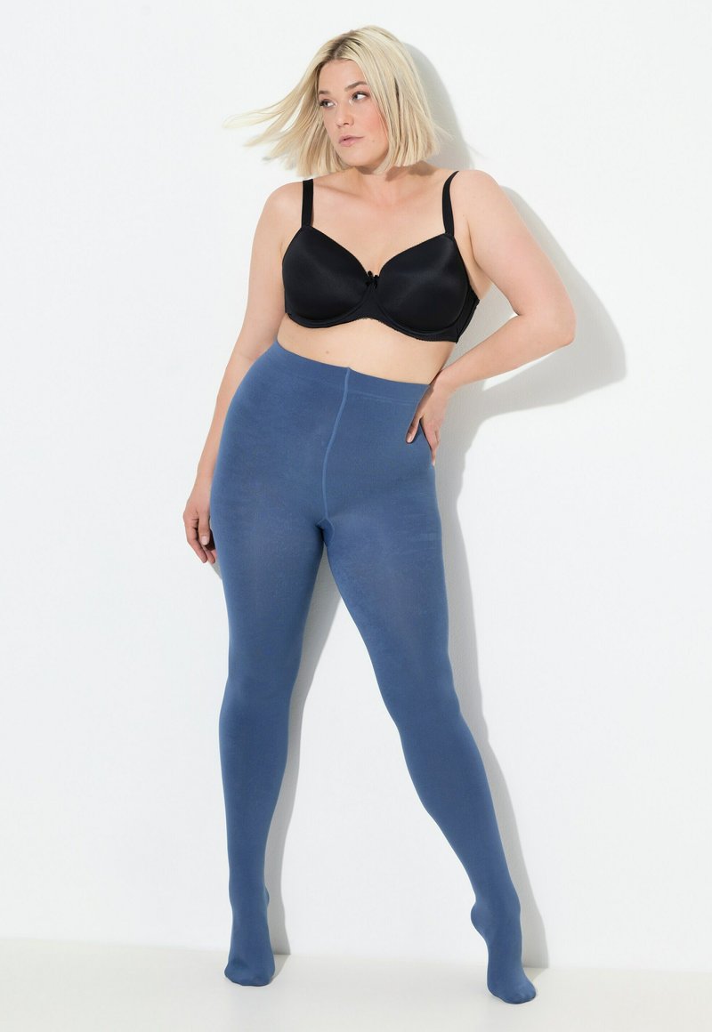 Frau mit kurzen blonden Haaren, die einen schwarzen BH und hochgeschnittene blaue Leggings trägt, posiert vor einer weißen Wand und schaut zur Seite.