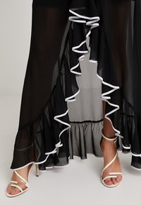 Schwarzer transparenter Rock mit weißem Rüschensaum und gestuftem Design. Inklusive weißer Sandaletten mit minimalistischen Details.